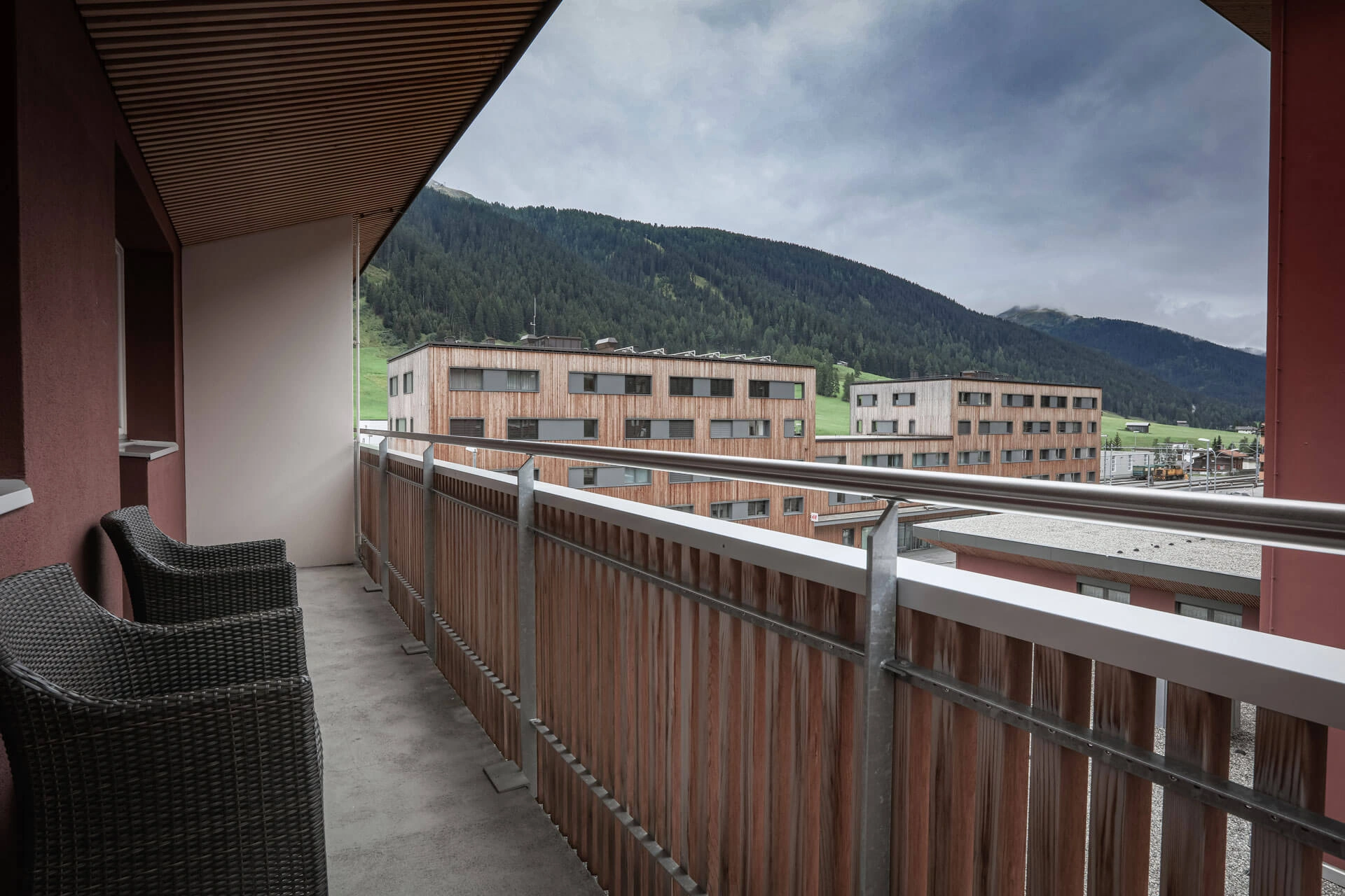 Überdachter Balkon mit zwei Korbsesseln und Holzgeländer, mit Blick auf moderne Gebäude und bewaldete Berghänge unter bewölktem Himmel.