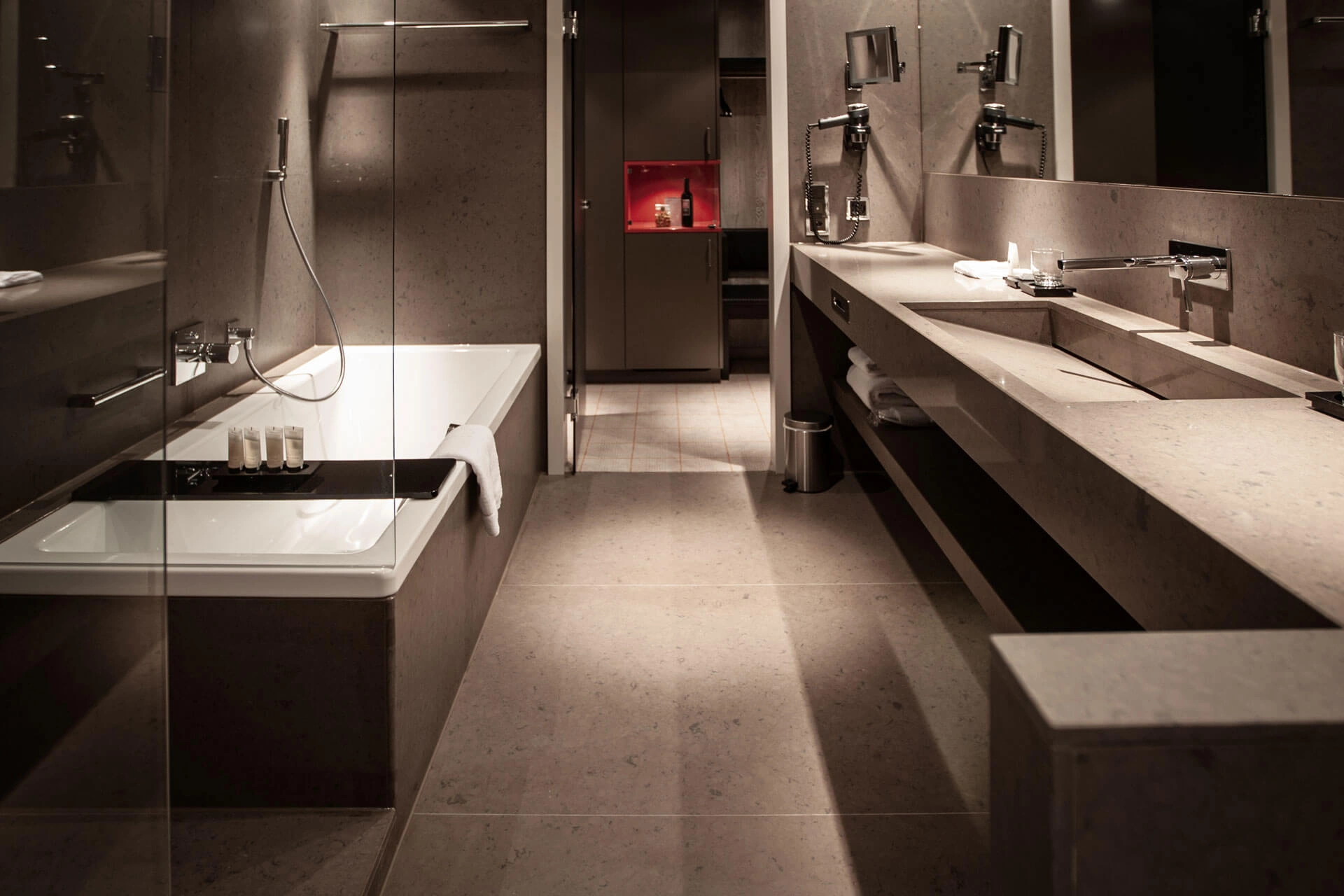 Modernes Badezimmer in warmen Grautönen mit Badewanne, Glasabtrennung, langem Waschtisch, großem Spiegel und dezent beleuchteten Pflegeprodukten.