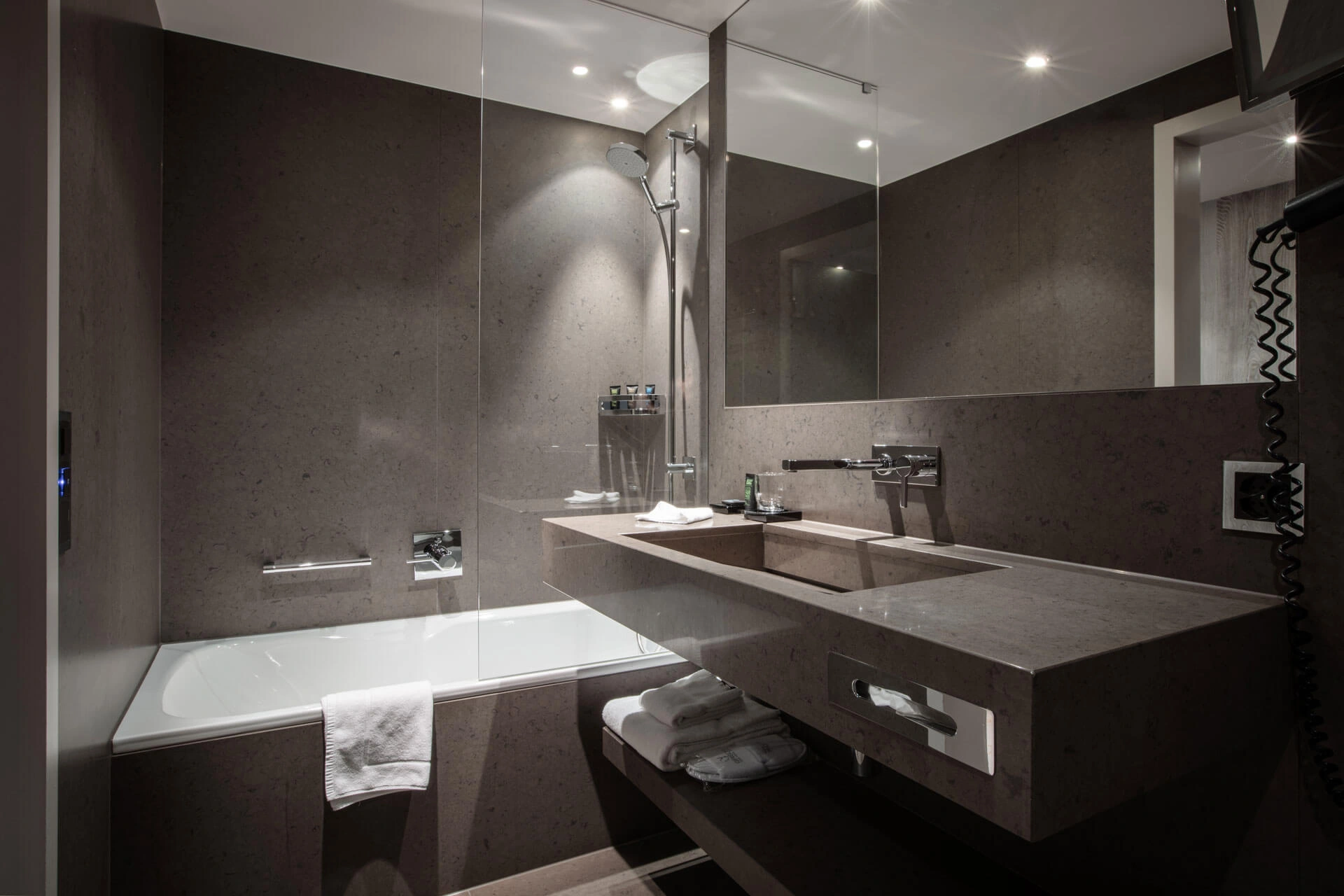 Modernes Badezimmer mit grauen Steinoberflächen, Badewanne mit Glasabtrennung, Regendusche, großem Spiegel und integriertem Waschtisch.