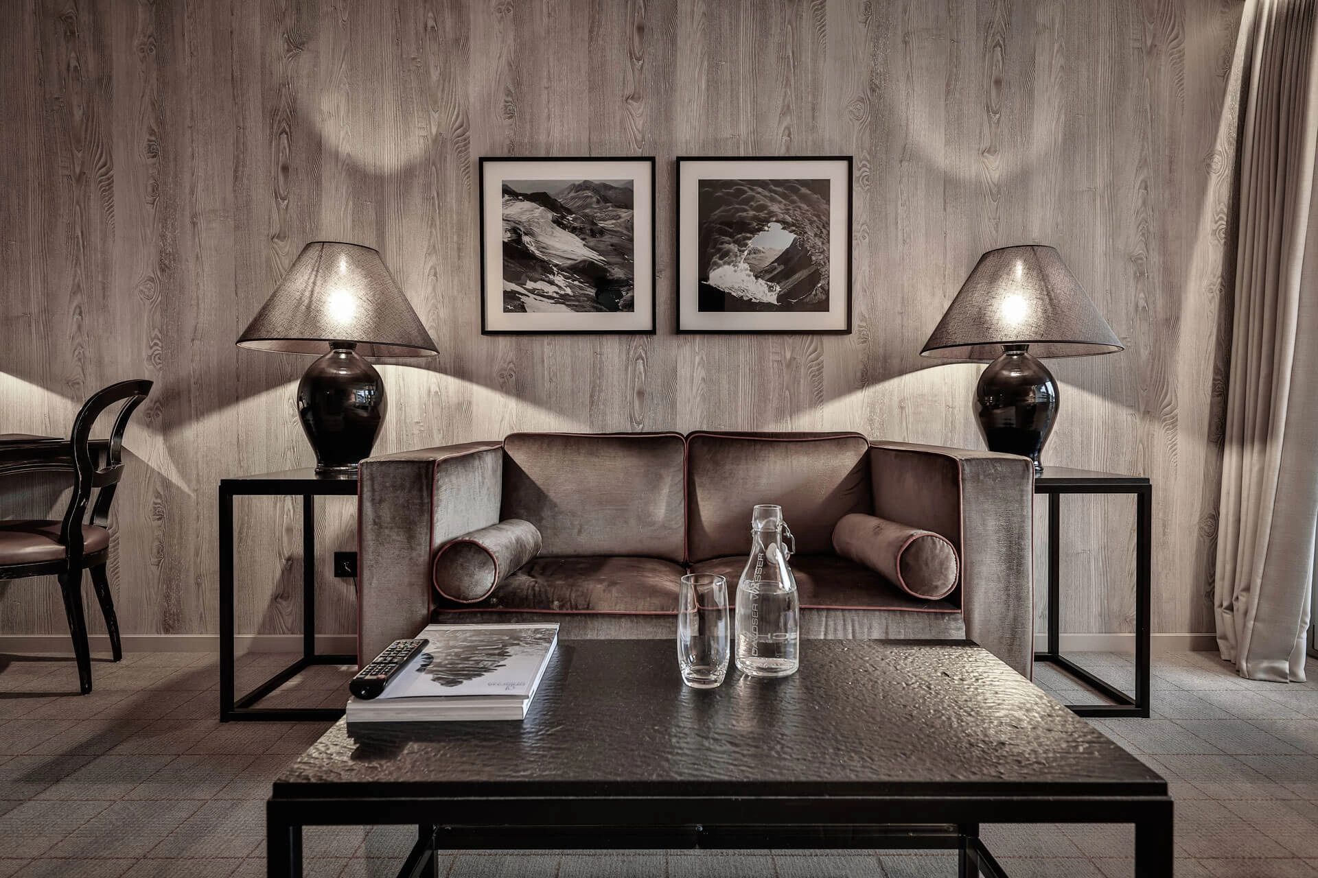 Elegante Sitzecke mit grauem Samtsofa, dunklem Couchtisch, zwei Tischlampen und gerahmten Schwarz-Weiß-Fotografien an der Holzwand.
