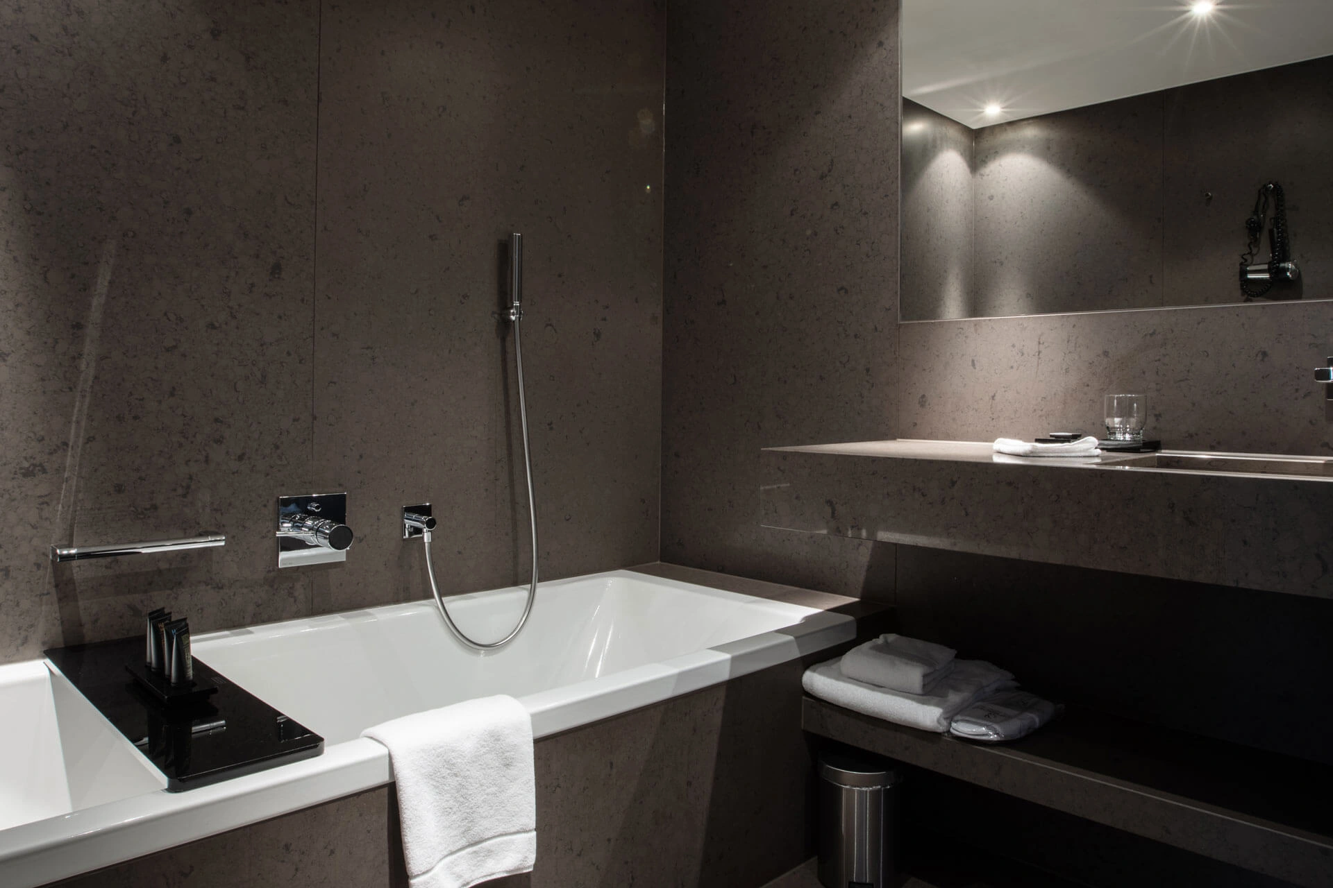 Modernes Badezimmer in dunklen Grautönen mit weißer Badewanne, Handbrause, großem Wandspiegel und Ablage mit Handtüchern.