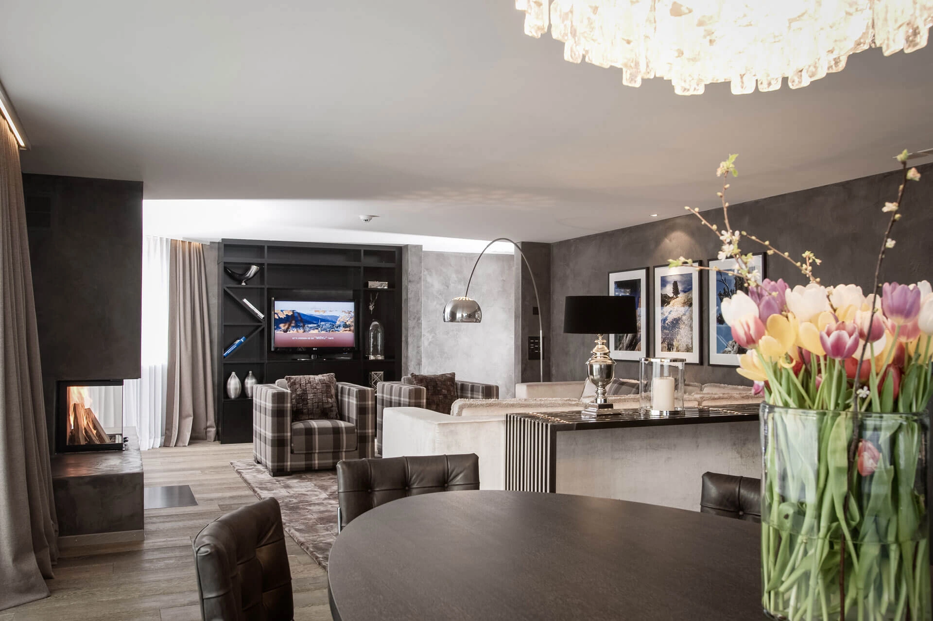 Eleganter Wohn- und Essbereich mit Kamin, Sesseln, Sofa und dunklen Möbeln. Ein großer Tulpenstrauß setzt einen farbigen Akzent.