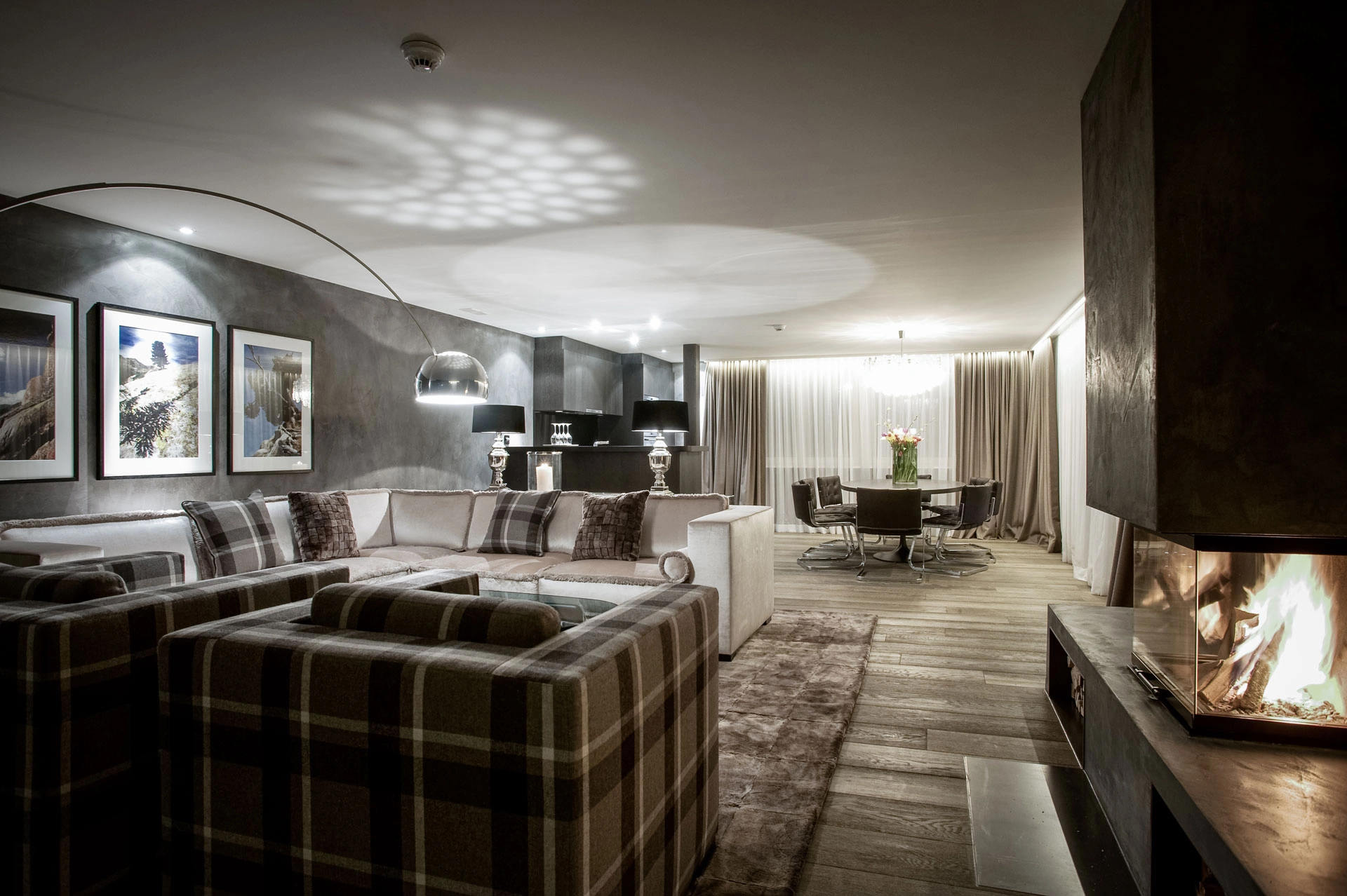 Großzügiger Wohn- und Essbereich mit hellem Sofa, karierten Sesseln, modernen Leuchten, gerahmten Bergmotiven und brennendem Kamin.