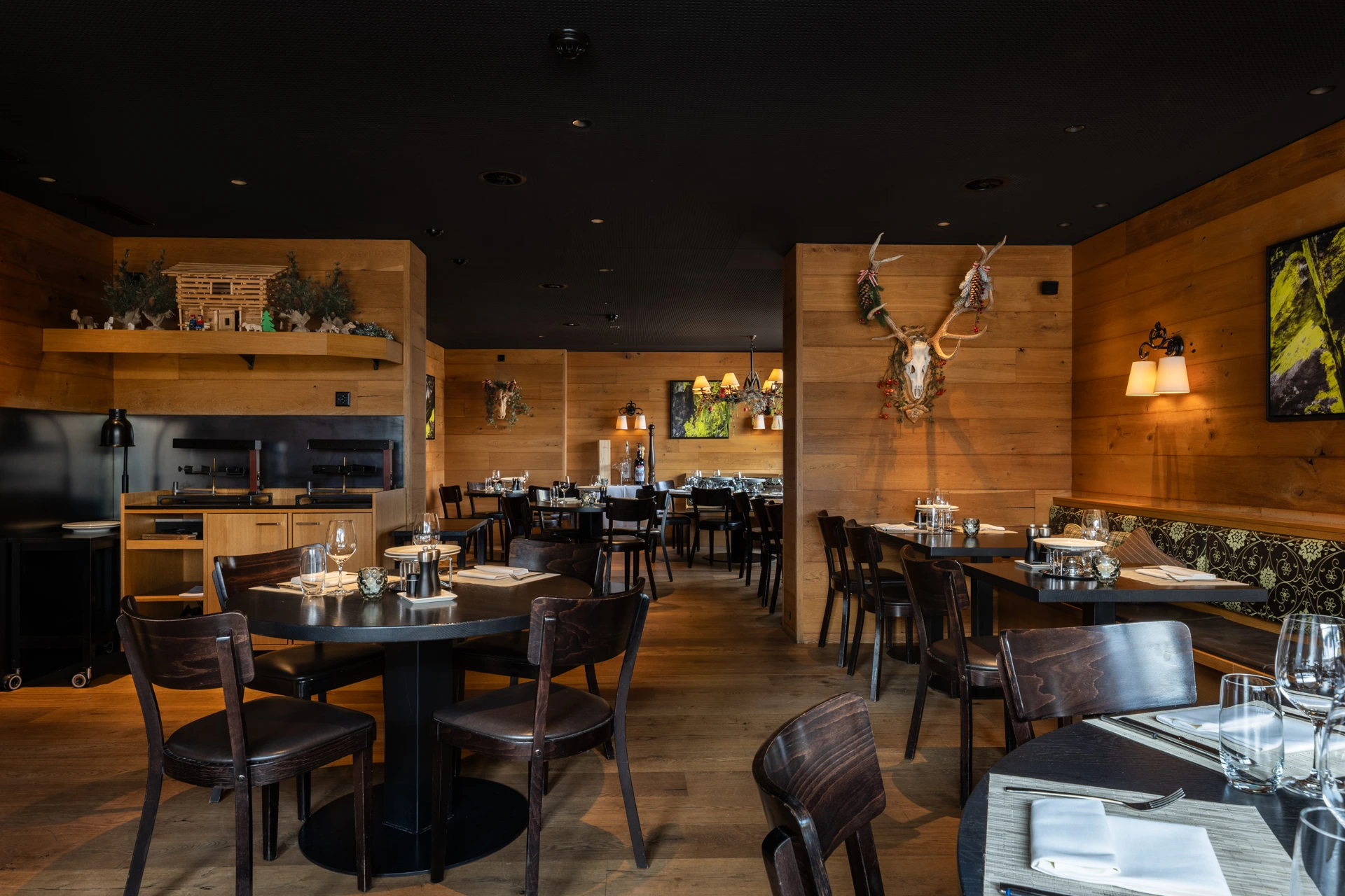 Gemütlicher Restaurantbereich mit warmen Holzwänden, dunkel gedeckten Tischen und Stühlen. Alpine Dekoration mit Geweih und Tiermotiven prägt den rustikalen Innenraum.