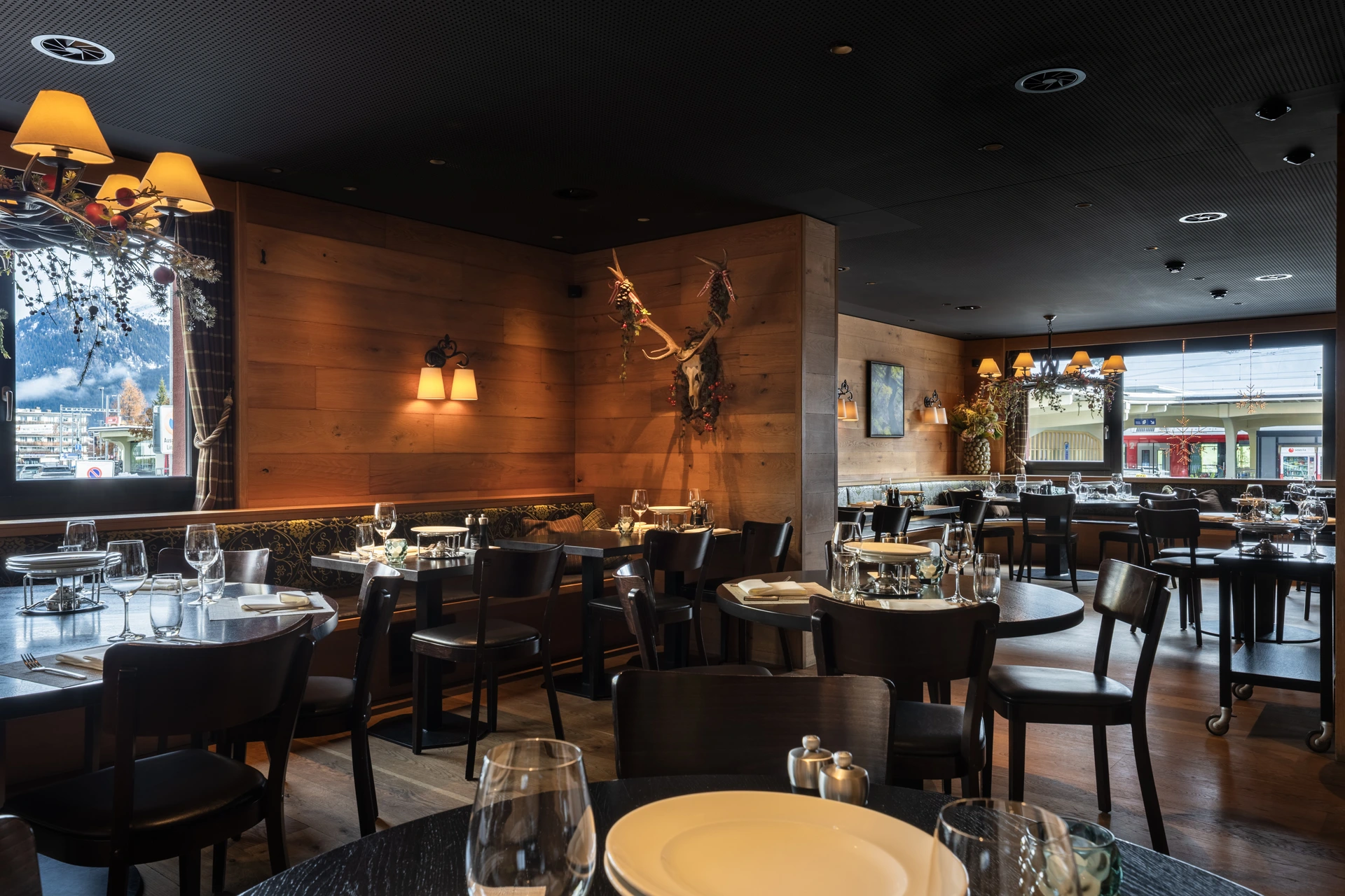 Stilvoller Restaurantbereich mit Holzwänden, dunklen Tischen und warmen Leuchten. Große Fenster geben den Blick auf die umliegende Berglandschaft und den Ort frei.