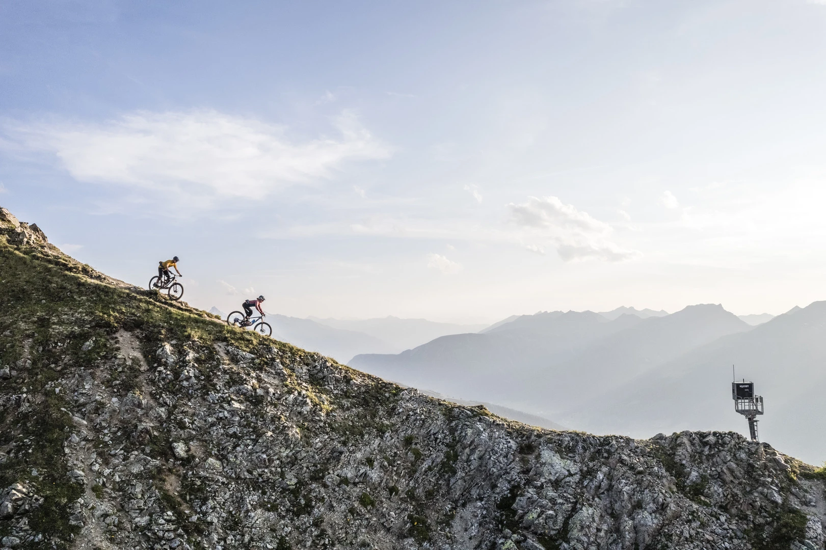 Zwei Mountainbiker auf einem Gratweg hoch über dem Tal im Abendlicht