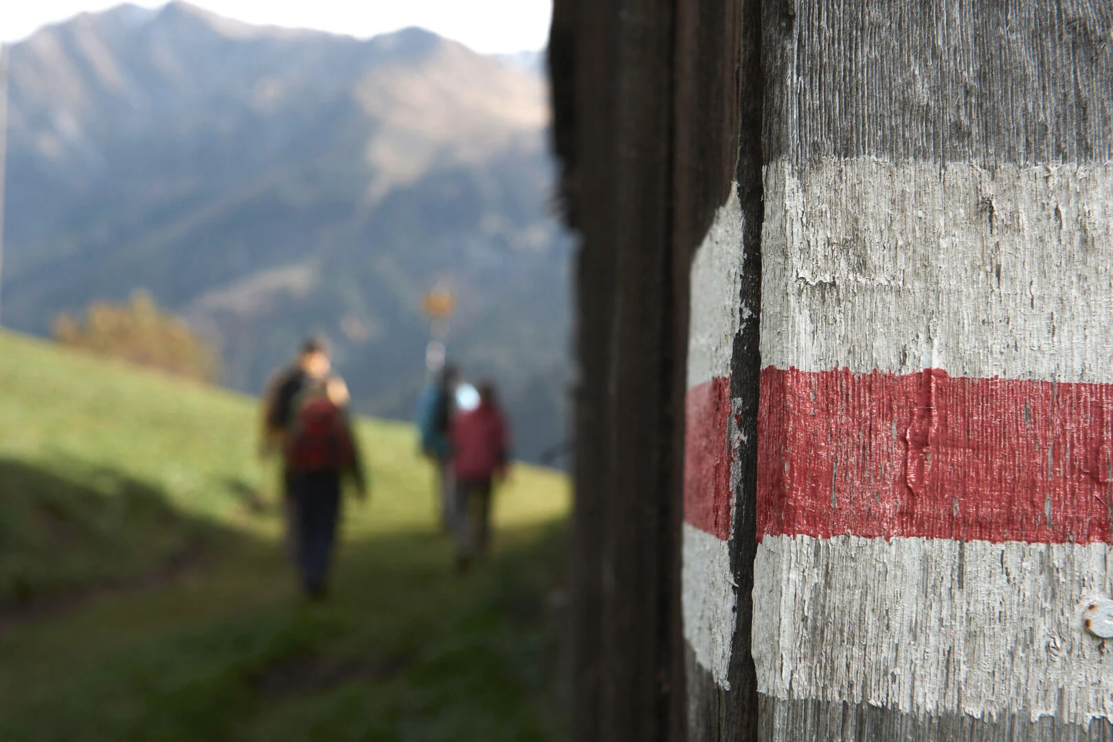 Rot-weisse Wanderwegmarkierung auf einem Holzpfosten, dahinter zwei Wanderer
