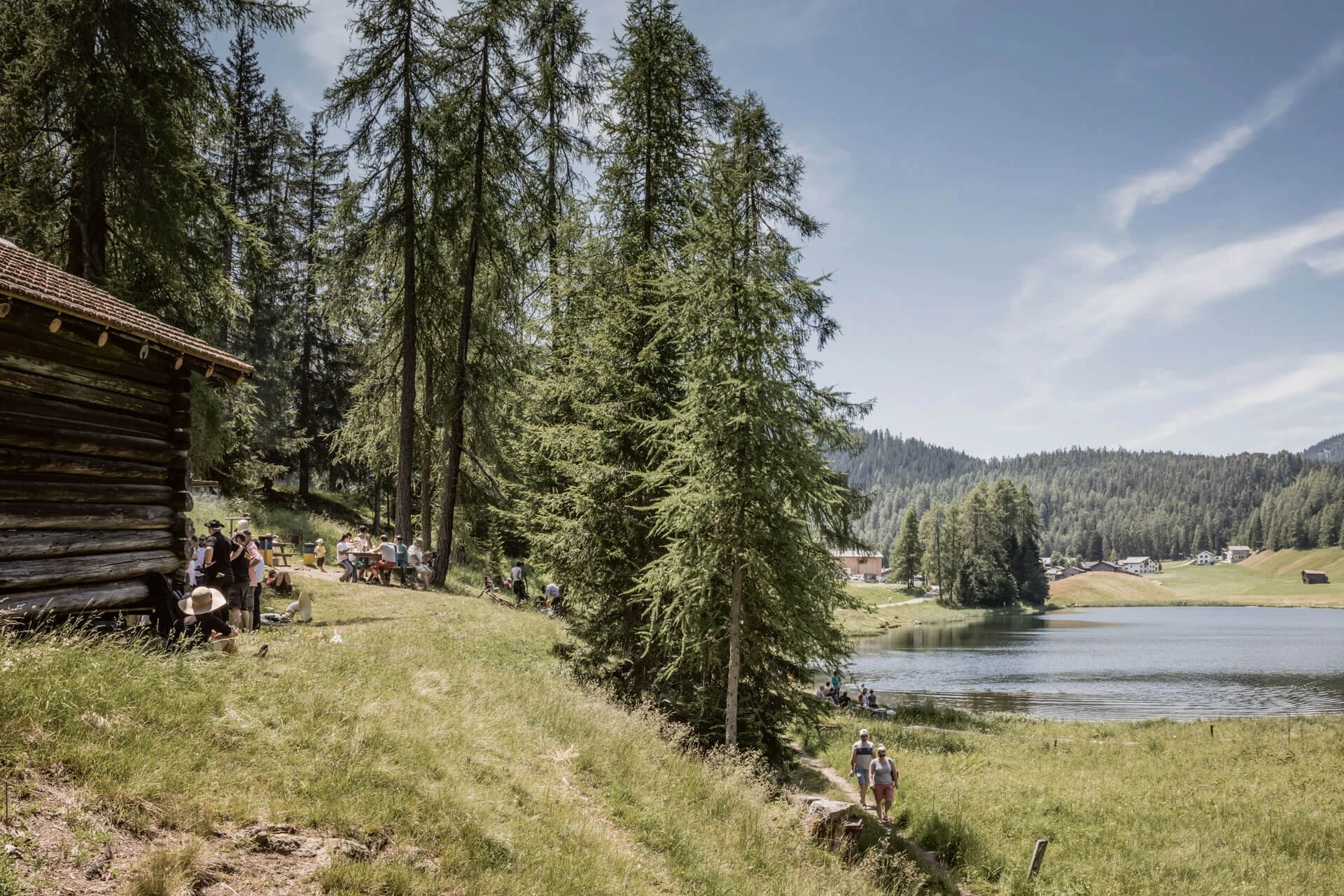 Gäste sitzen vor einem Blockhaus am Ufer eines Bergsees