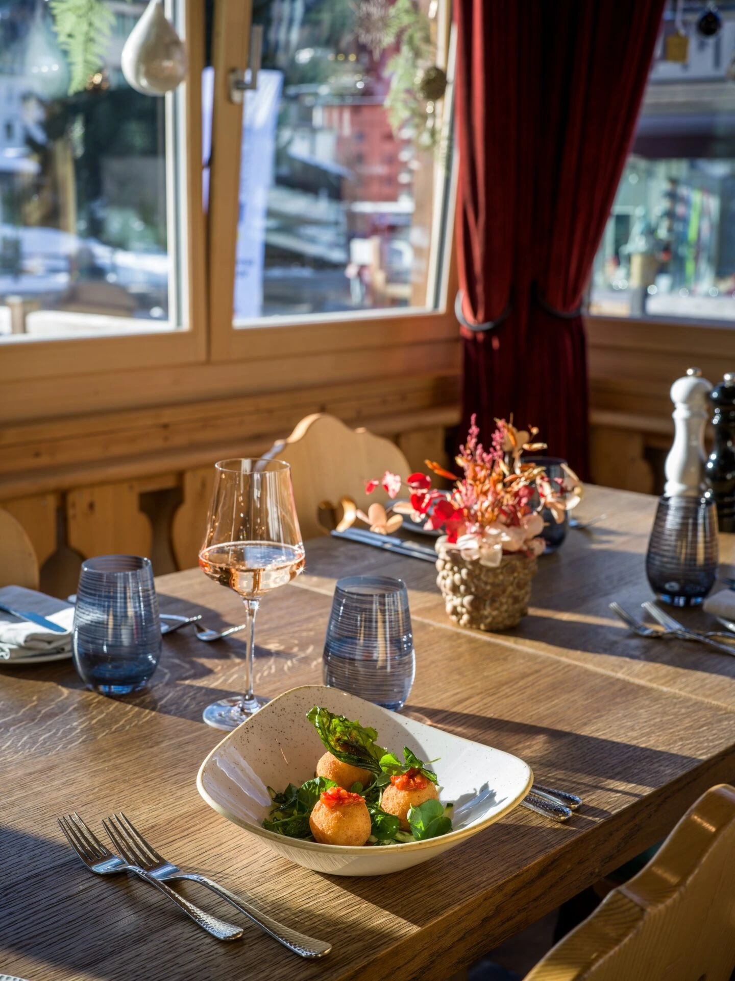 Sonnendurchfluteter Restauranttisch am Fenster mit stilvoll angerichteter Vorspeise, Weinglas und herbstlicher Tischdekoration.